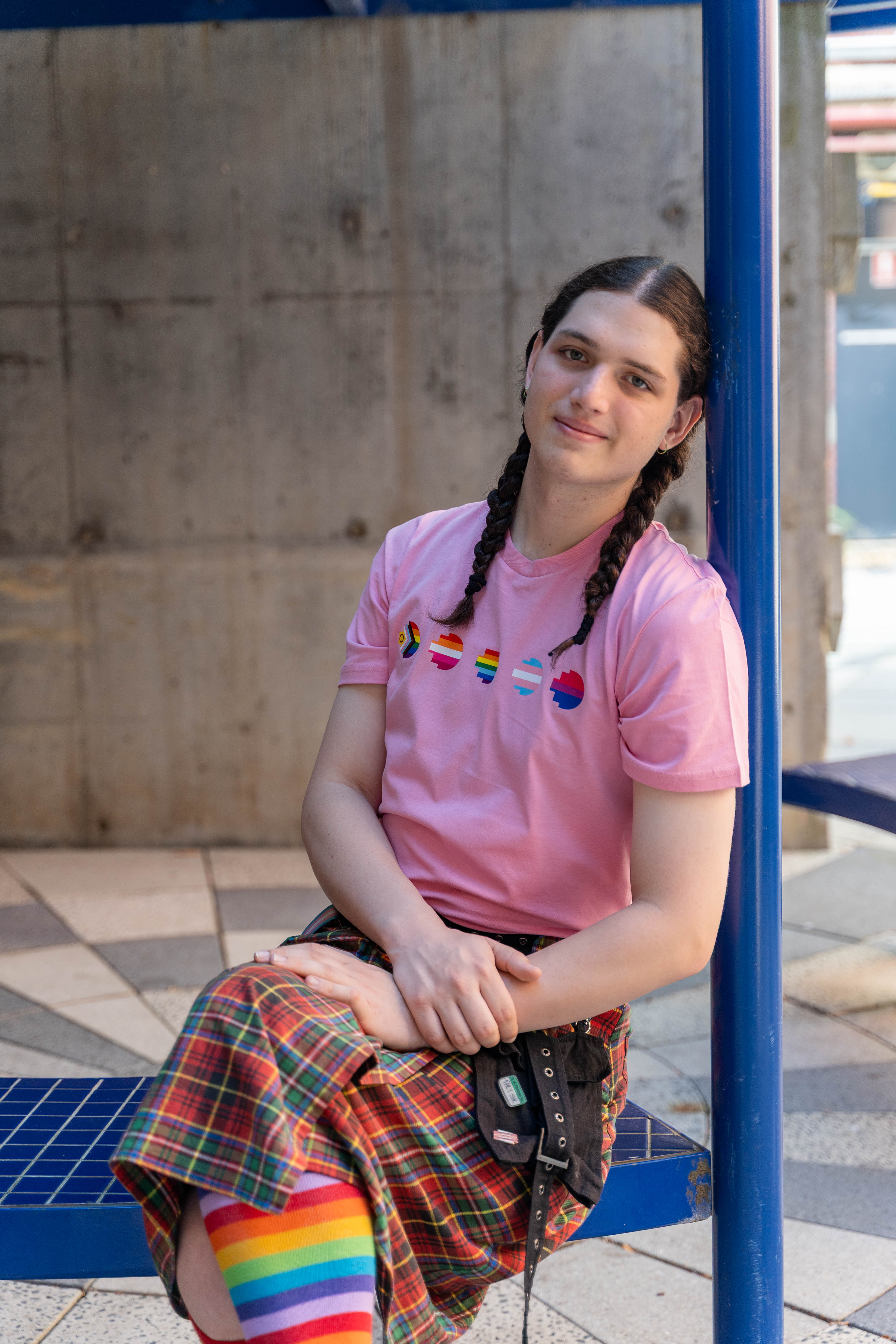 Model seated wearing the limited edition pink Pride Unity Tee from RMIT Store, complemented by a plaid skirt and rainbow socks, highlighting pride flag designs on the chest.